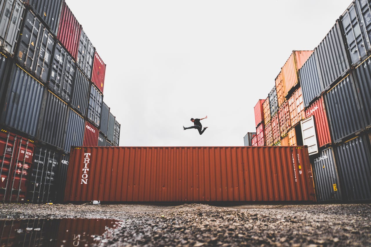 A person leaps dramatically above stacked shipping containers, showcasing freedom and industrial energy.